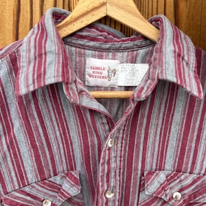 May include: A button-down shirt with vertical stripes in burgundy and gray. The shirt has two chest pockets and a collar. The label reads "Saddle King Western". The shirt is hanging on a wooden hanger.