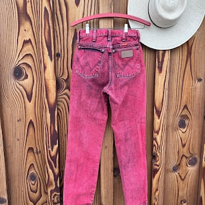 May include: A pair of acid-wash pink Wrangler jeans hangs on a pink hanger against a wooden background. The jeans have a classic five-pocket design with the Wrangler logo on the back pocket. A white straw hat rests on the wooden surface.