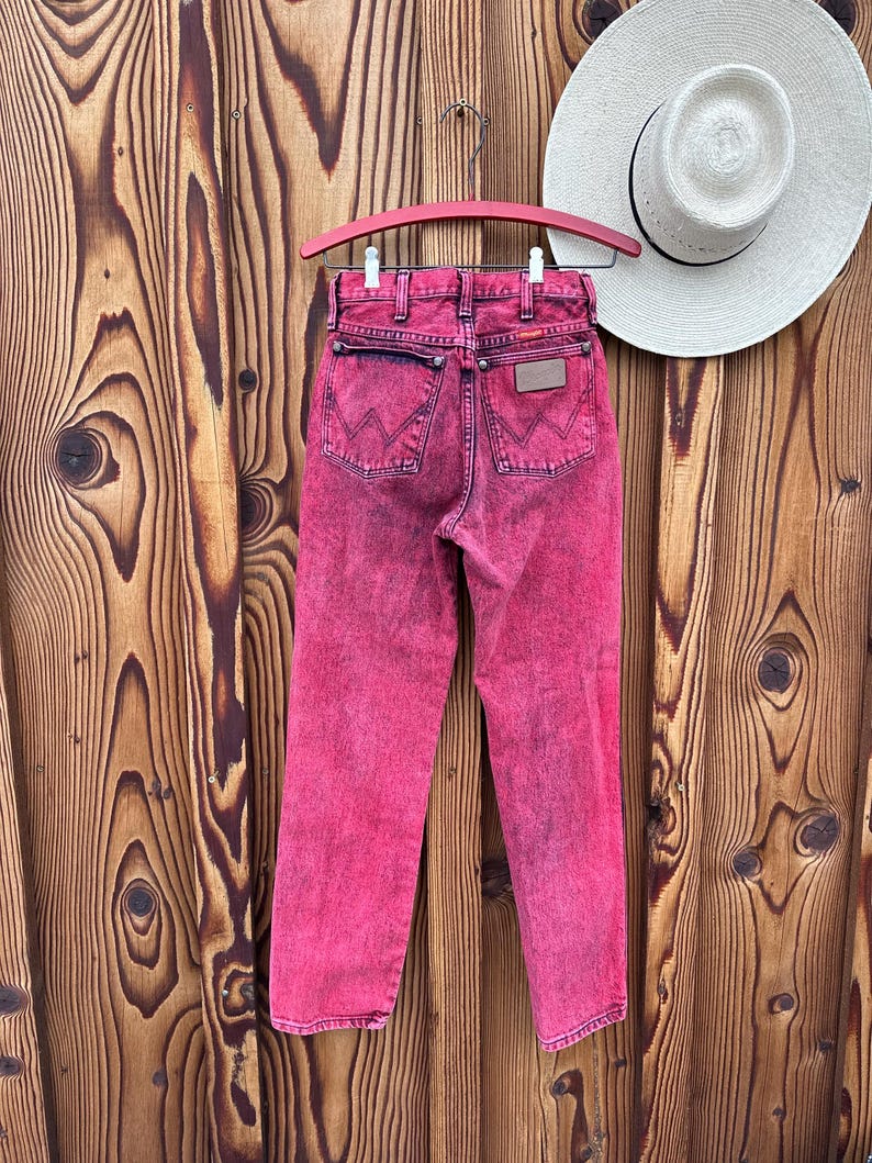 May include: A pair of acid-wash pink Wrangler jeans hangs on a pink hanger against a wooden background. The jeans have a classic five-pocket design with the Wrangler logo on the back pocket. A white straw hat rests on the wooden surface.