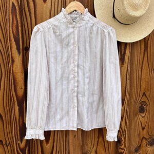May include: A vintage white blouse with vertical pink stripes, a high ruffled collar, and long sleeves. The blouse is buttoned down the front and has ruffled cuffs. A straw hat with a black band hangs in the background.