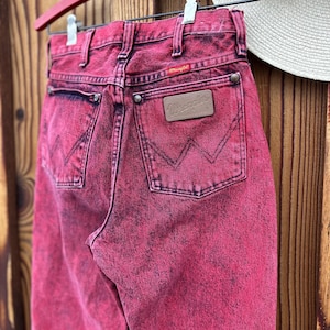May include: A pair of vintage Wrangler jeans in a vibrant pink hue, hanging on a red hanger. The jeans feature classic five-pocket styling, a leather patch, and the Wrangler logo. The denim has a faded, acid-wash effect.
