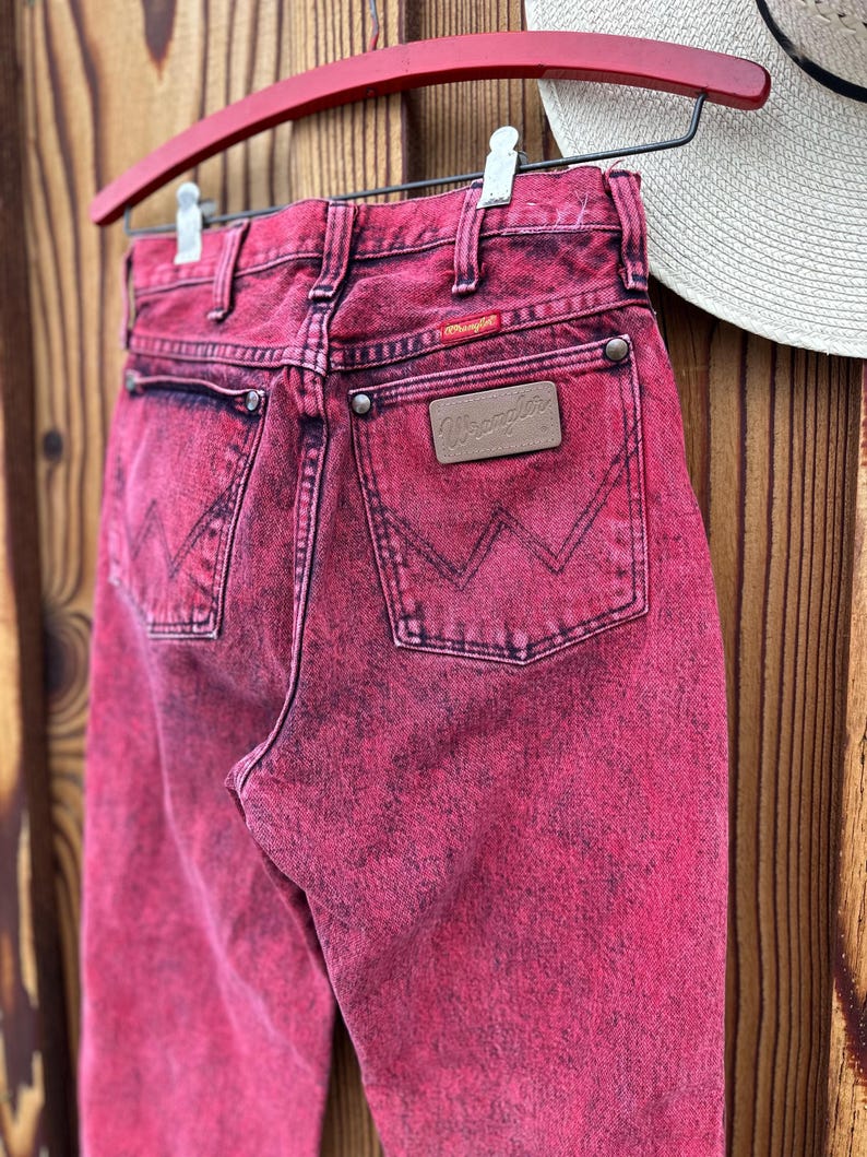 May include: A pair of vintage Wrangler jeans in a vibrant pink hue, hanging on a red hanger. The jeans feature classic five-pocket styling, a leather patch, and the Wrangler logo. The denim has a faded, acid-wash effect.