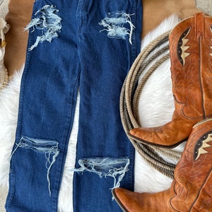 May include: Distressed blue denim jeans with multiple rips and tears. The jeans are laid out on a white fuzzy surface with a pair of brown cowboy boots and a straw hat.