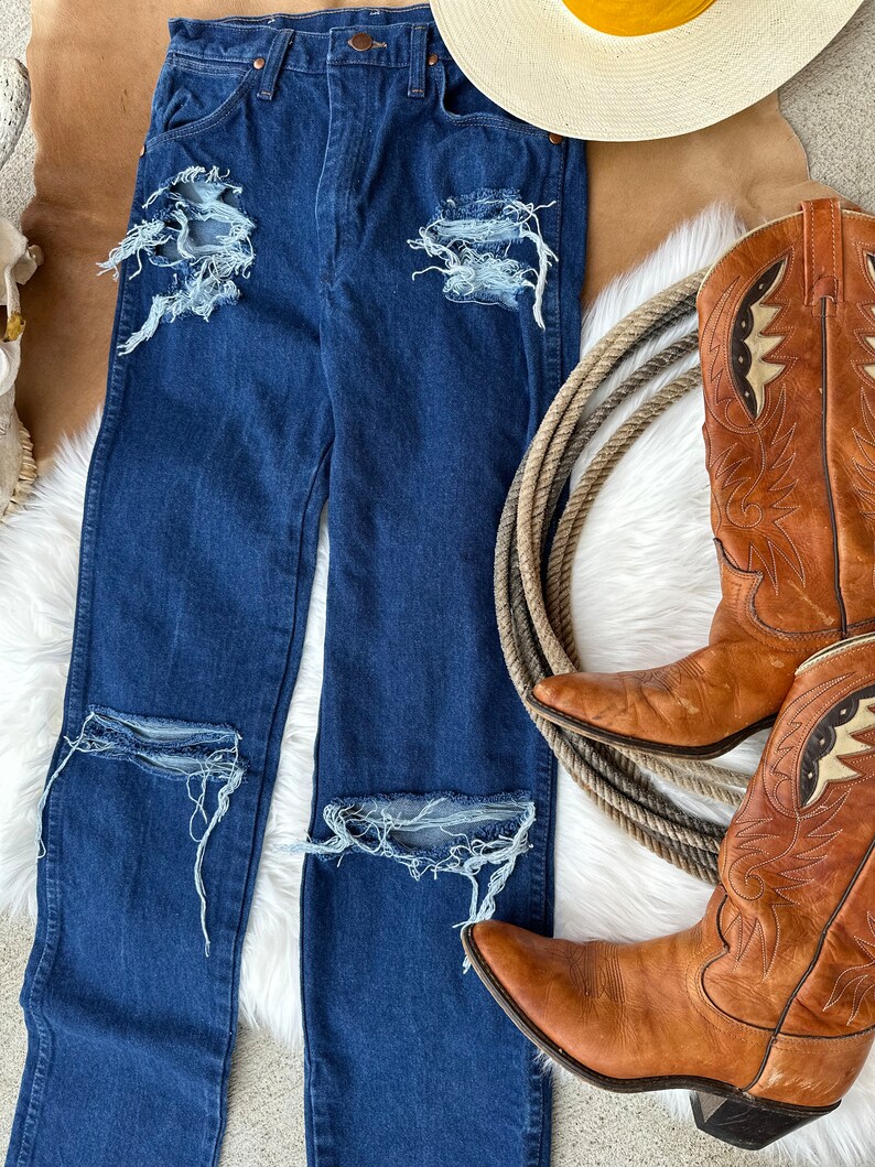 May include: Distressed blue denim jeans with multiple rips and tears. The jeans are laid out on a white fuzzy surface with a pair of brown cowboy boots and a straw hat.