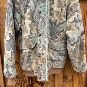 May include: Camouflage jacket with a leaf pattern in shades of brown, green, and gray. The jacket has a button-up front, two front pockets, and brown ribbed cuffs and hem. The jacket is made of a durable material.