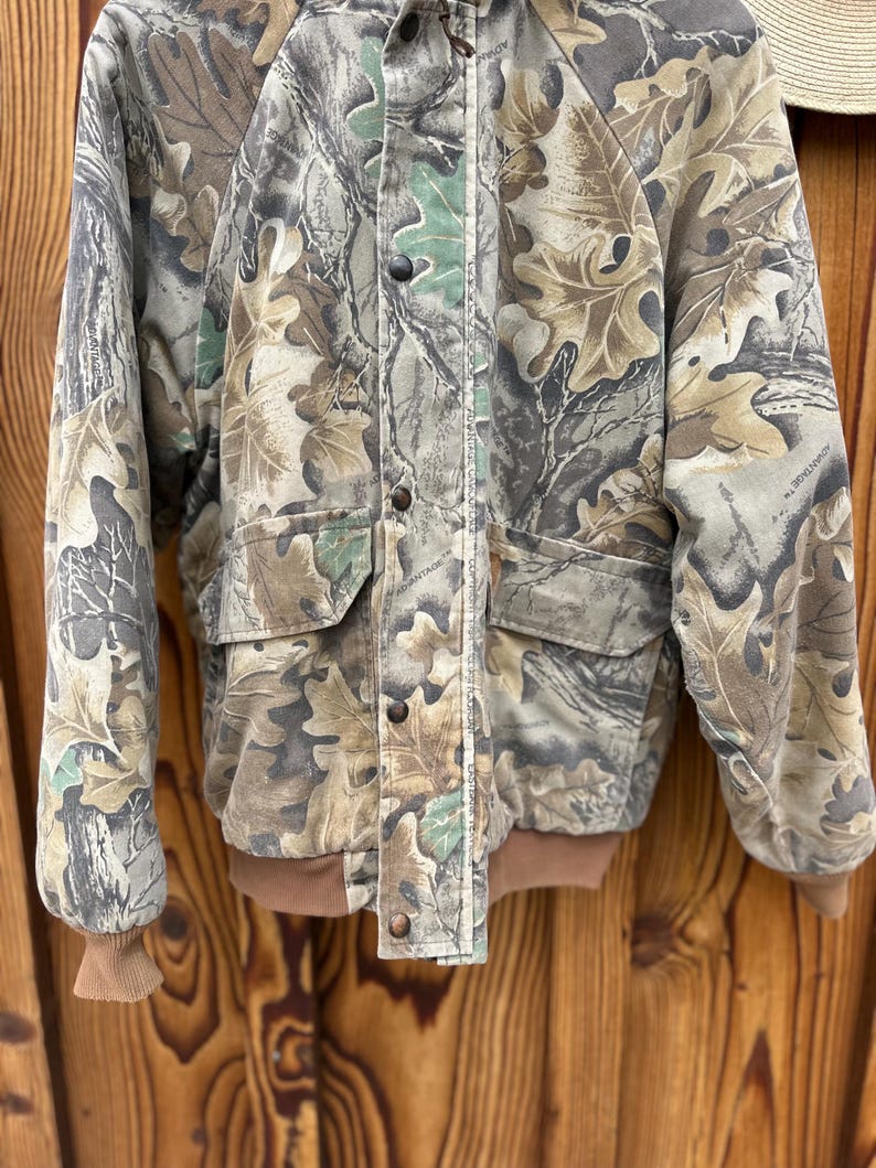 May include: Camouflage jacket with a leaf pattern in shades of brown, green, and gray. The jacket has a button-up front, two front pockets, and brown ribbed cuffs and hem. The jacket is made of a durable material.