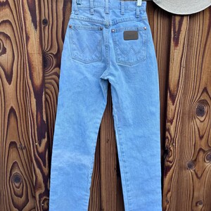 May include: Light blue denim jeans with a classic five-pocket design, hanging on a clear hanger. The jeans feature a brown leather patch on the back pocket and a straight leg cut. The Wrangler brand is visible on the patch.