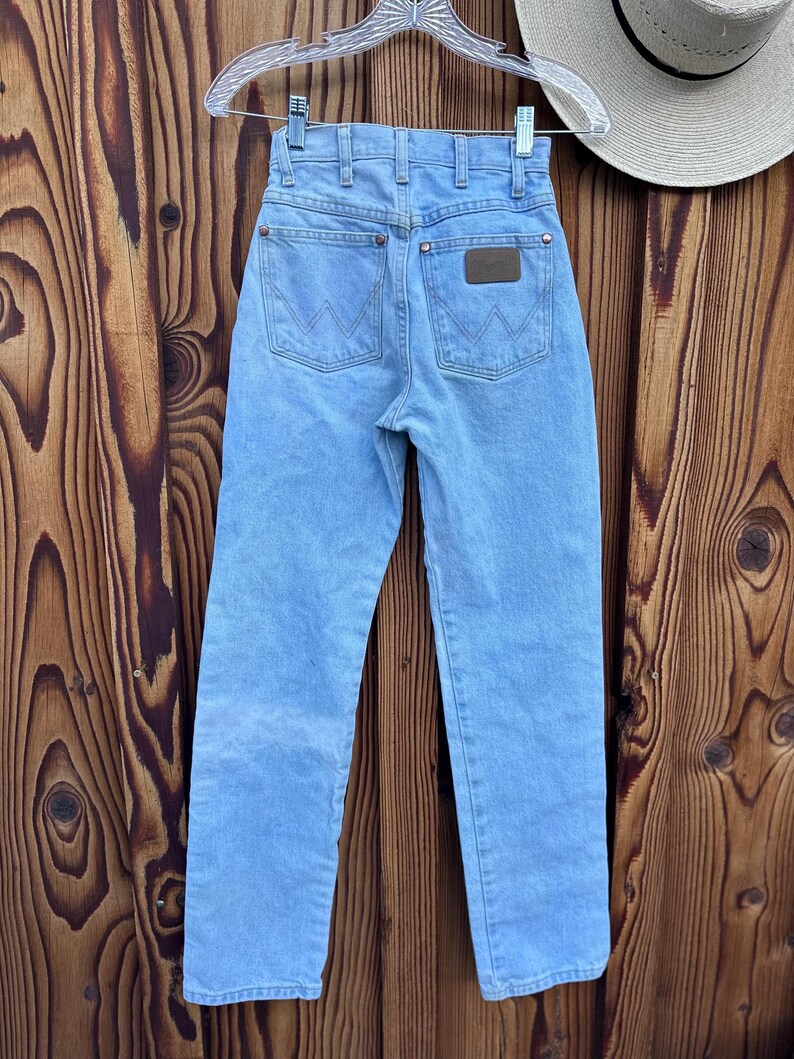 May include: Light blue denim jeans with a classic five-pocket design, hanging on a clear hanger. The jeans feature a brown leather patch on the back pocket and a straight leg cut. The Wrangler brand is visible on the patch.