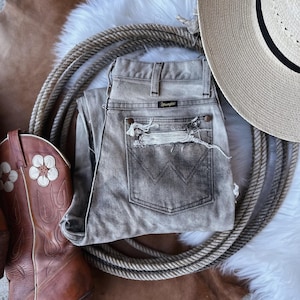 May include: Distressed light gray denim jeans with a brown leather belt loop and a Wrangler label. The jeans are lying on a white fur rug with a brown leather background and a straw cowboy hat.