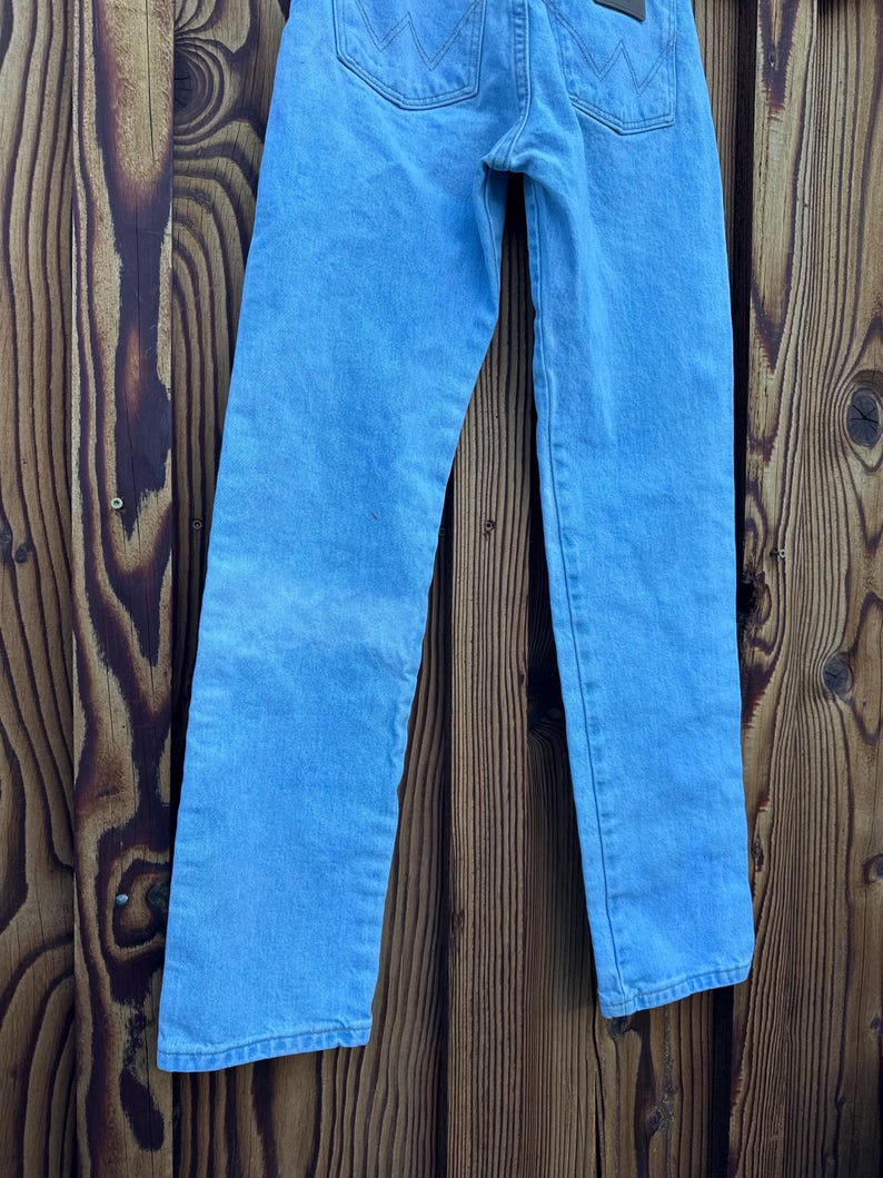 May include: Light blue denim jeans with a classic five-pocket design. The jeans have a straight leg cut and a faded wash, with visible stitching details. The Wrangler brand label is visible on the back pocket.