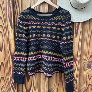May include: A black knit sweater with a colorful geometric pattern in pink, gold, and gray. The sweater is hanging on a wooden hanger against a wooden background. A straw hat is partially visible.