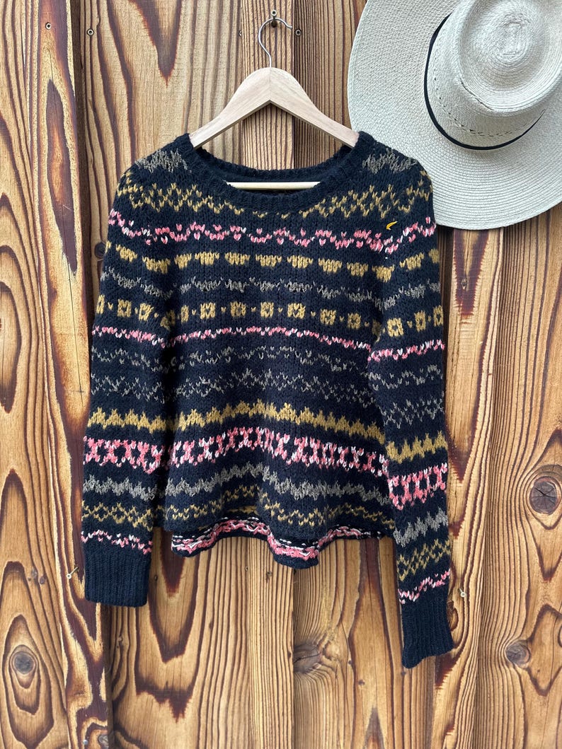 May include: A black knit sweater with a colorful geometric pattern in pink, gold, and gray. The sweater is hanging on a wooden hanger against a wooden background. A straw hat is partially visible.