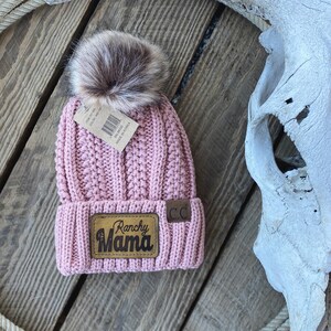 May include: A pink knitted beanie with a faux fur pom pom and a brown leather patch that says "Ranchy Mama".