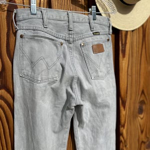 Pu&ograve; includere: Jeans Wrangler grigio chiaro con classico design a cinque tasche, toppa in pelle marrone e logo Wrangler. I jeans sono appesi a una gruccia trasparente su sfondo in legno. Si vede un cappello di paglia.