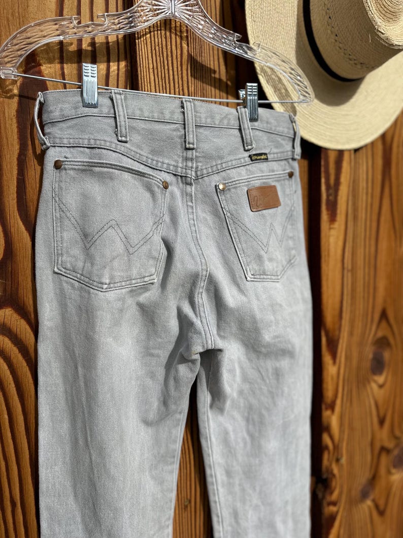 Pu&ograve; includere: Jeans Wrangler grigio chiaro con classico design a cinque tasche, toppa in pelle marrone e logo Wrangler. I jeans sono appesi a una gruccia trasparente su sfondo in legno. Si vede un cappello di paglia.