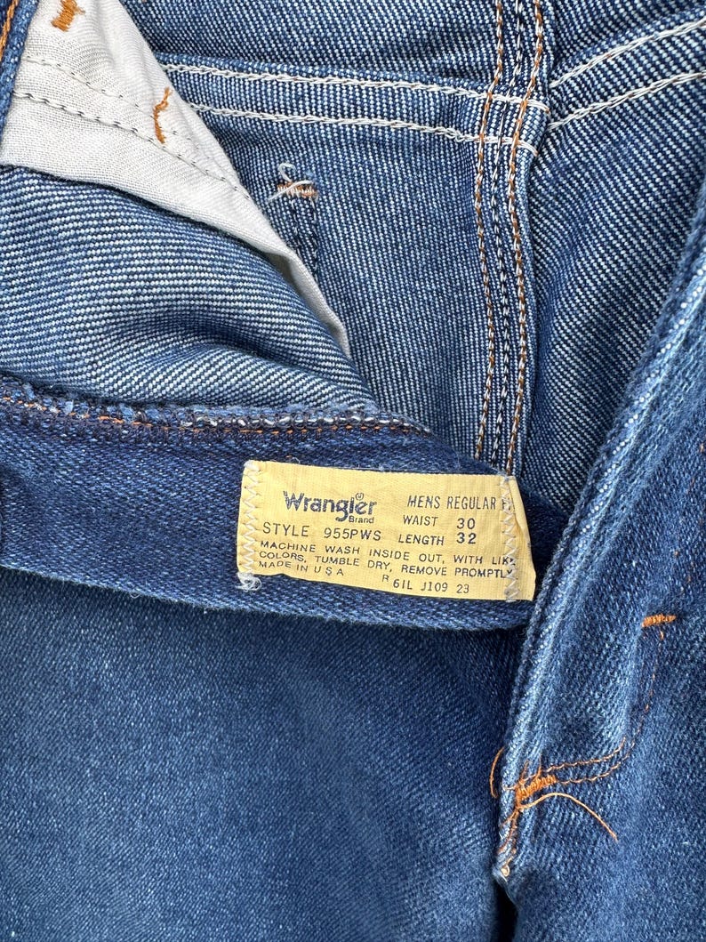 Puede incluir: Un vaquero azul de mezclilla Wrangler para hombre, de corte regular, con una etiqueta de cuero marr&oacute;n. La etiqueta tiene el texto "Wrangler Brand", "Mens Regular Fit", "Style 955PWS", "Waist 30", "Length 32", "Machine Wash Inside Out, With Like Colors, Tumble Dry, Remove Promptly", "Made in USA", "R6IL J109 23".