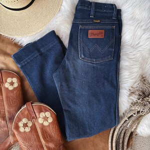 Puede incluir: Vaqueros de mezclilla azul oscuro con un parche de cuero marr&oacute;n Wrangler en el bolsillo trasero. Los vaqueros tienen una pierna recta y una cintura alta.