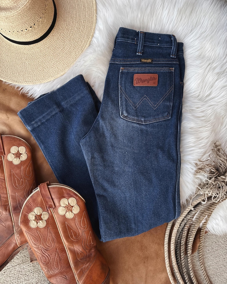 Puede incluir: Vaqueros de mezclilla azul oscuro con un parche de cuero marr&oacute;n Wrangler en el bolsillo trasero. Los vaqueros tienen una pierna recta y una cintura alta.