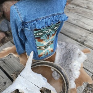 K&ouml;nnte beinhalten: Eine blaue Jeansjacke mit einem Stoffpanel im S&uuml;dwest-Stil auf der R&uuml;ckseite. Die Jacke hat einen R&uuml;schenbesatz um den r&uuml;ckw&auml;rtigen Halsausschnitt.