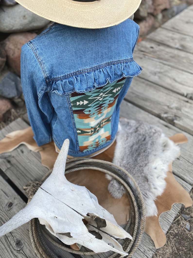 K&ouml;nnte beinhalten: Eine blaue Jeansjacke mit einem Stoffpanel im S&uuml;dwest-Stil auf der R&uuml;ckseite. Die Jacke hat einen R&uuml;schenbesatz um den r&uuml;ckw&auml;rtigen Halsausschnitt.