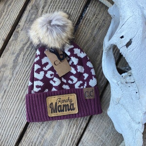 May include: A burgundy knit beanie with a faux fur pom-pom and a white leopard print pattern. The hat features a brown leather patch that reads "Ranchy Mama". A small tag with "C.C" is attached.
