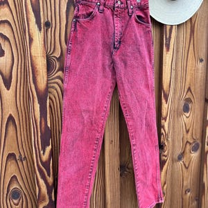 May include: A pair of acid-wash pink denim jeans hanging on a red wooden hanger. The jeans have a classic five-pocket design and a straight leg. A white hat with a black band hangs in the background.