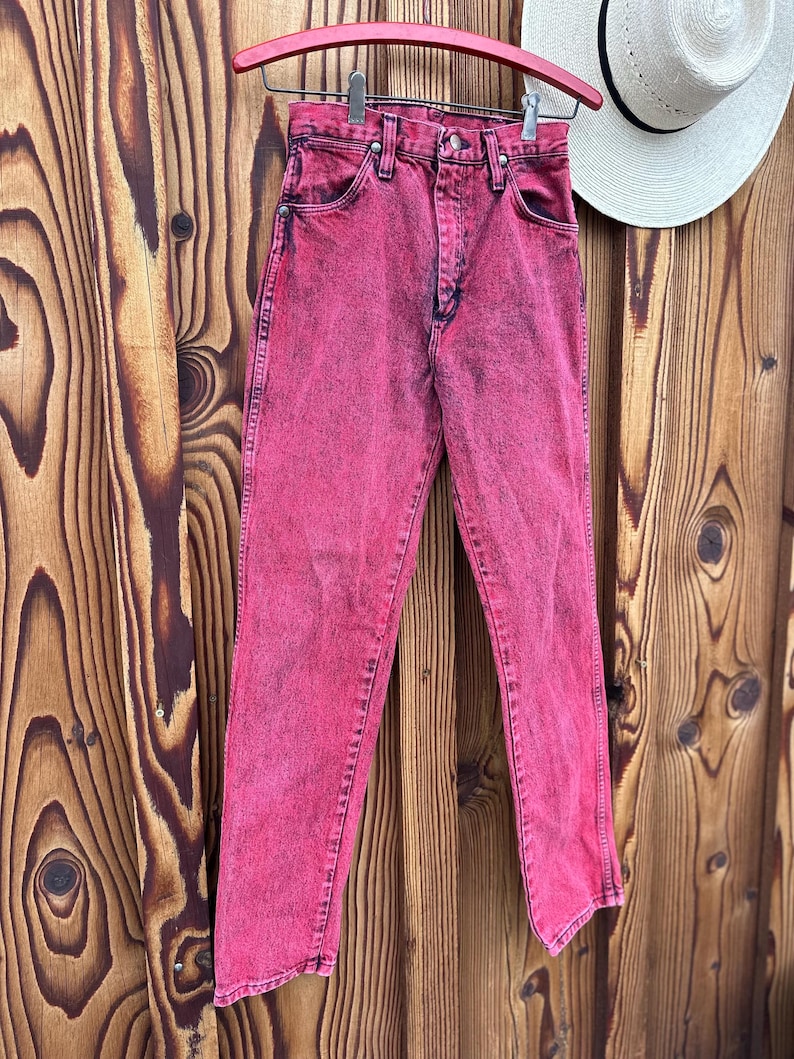 May include: A pair of acid-wash pink denim jeans hanging on a red wooden hanger. The jeans have a classic five-pocket design and a straight leg. A white hat with a black band hangs in the background.