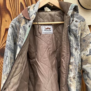 May include: Camouflage hooded jacket with a brown quilted lining. The jacket features a brown collar and trim, and a label with a duck logo. The jacket is hanging on a wooden hanger against a wooden background.