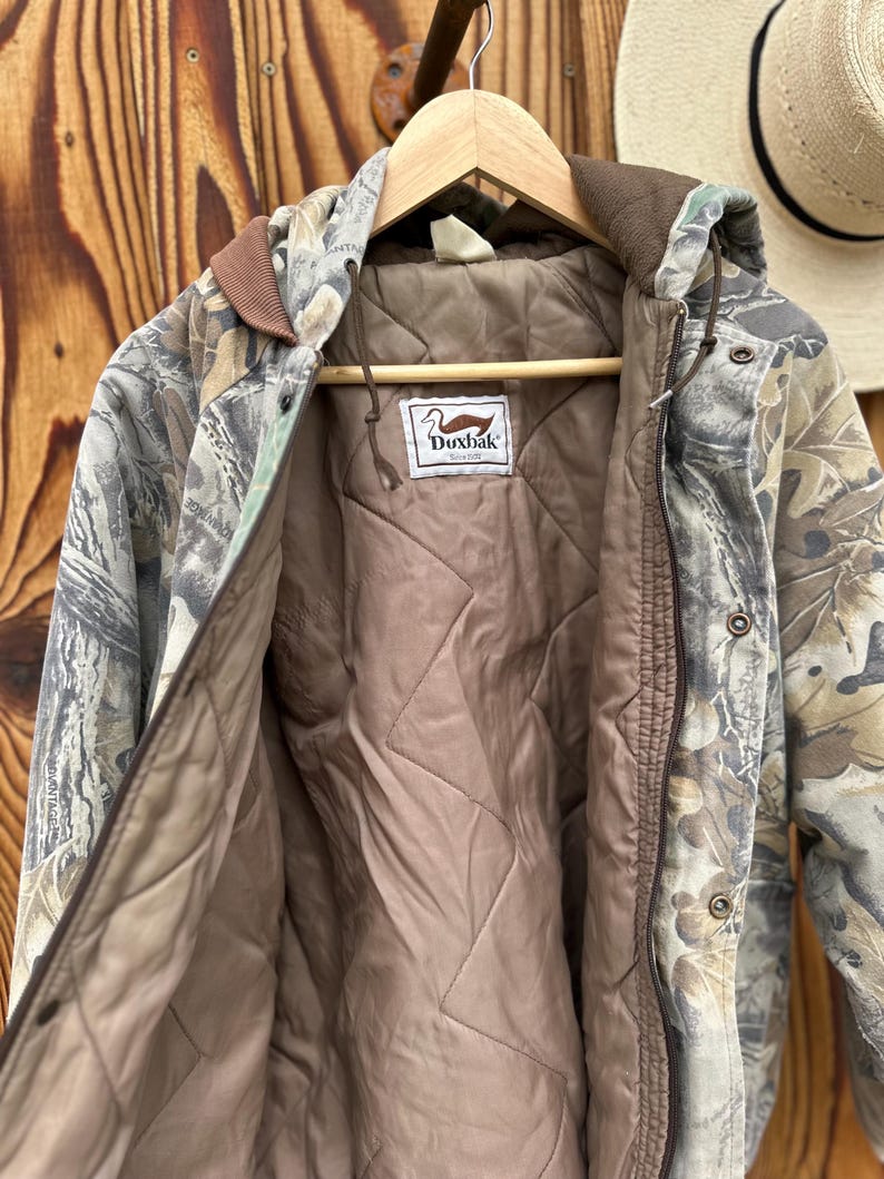 May include: Camouflage hooded jacket with a brown quilted lining. The jacket features a brown collar and trim, and a label with a duck logo. The jacket is hanging on a wooden hanger against a wooden background.