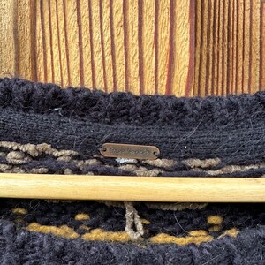 May include: Close-up of a black knit sweater with a brown and tan pattern. The sweater is hanging on a wooden hanger against a wooden background. A small metal tag with the words "Free People" is visible.
