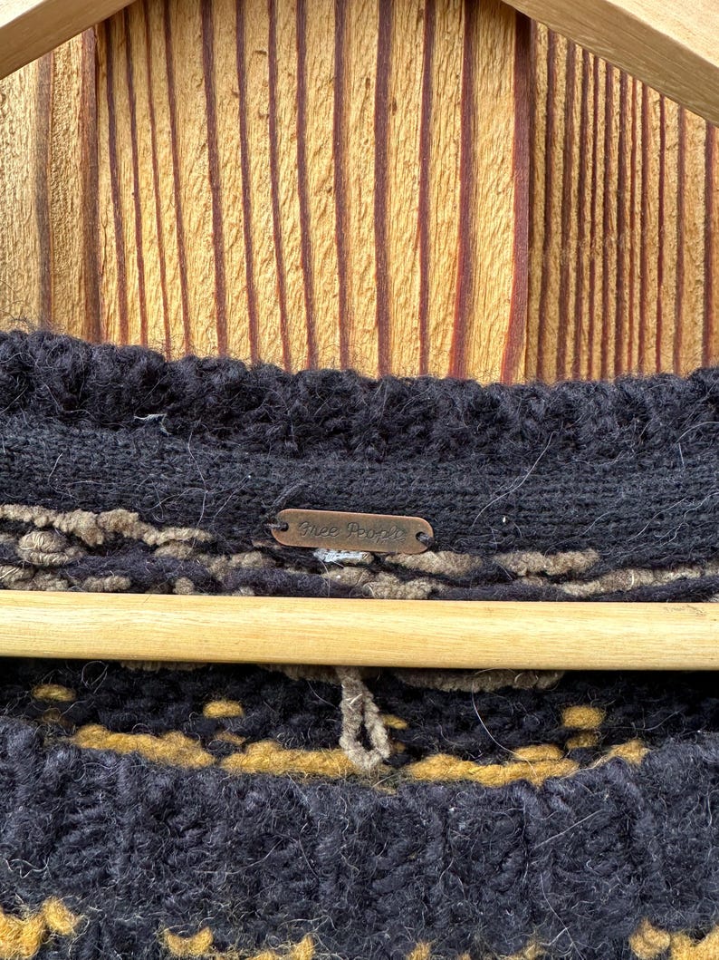 May include: Close-up of a black knit sweater with a brown and tan pattern. The sweater is hanging on a wooden hanger against a wooden background. A small metal tag with the words "Free People" is visible.