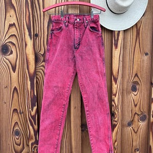 May include: A pair of acid-wash pink denim jeans hangs on a pink hanger against a wooden background. The jeans have a classic five-pocket design and a button and zipper closure. A white straw hat with a black band rests on the wooden surface.