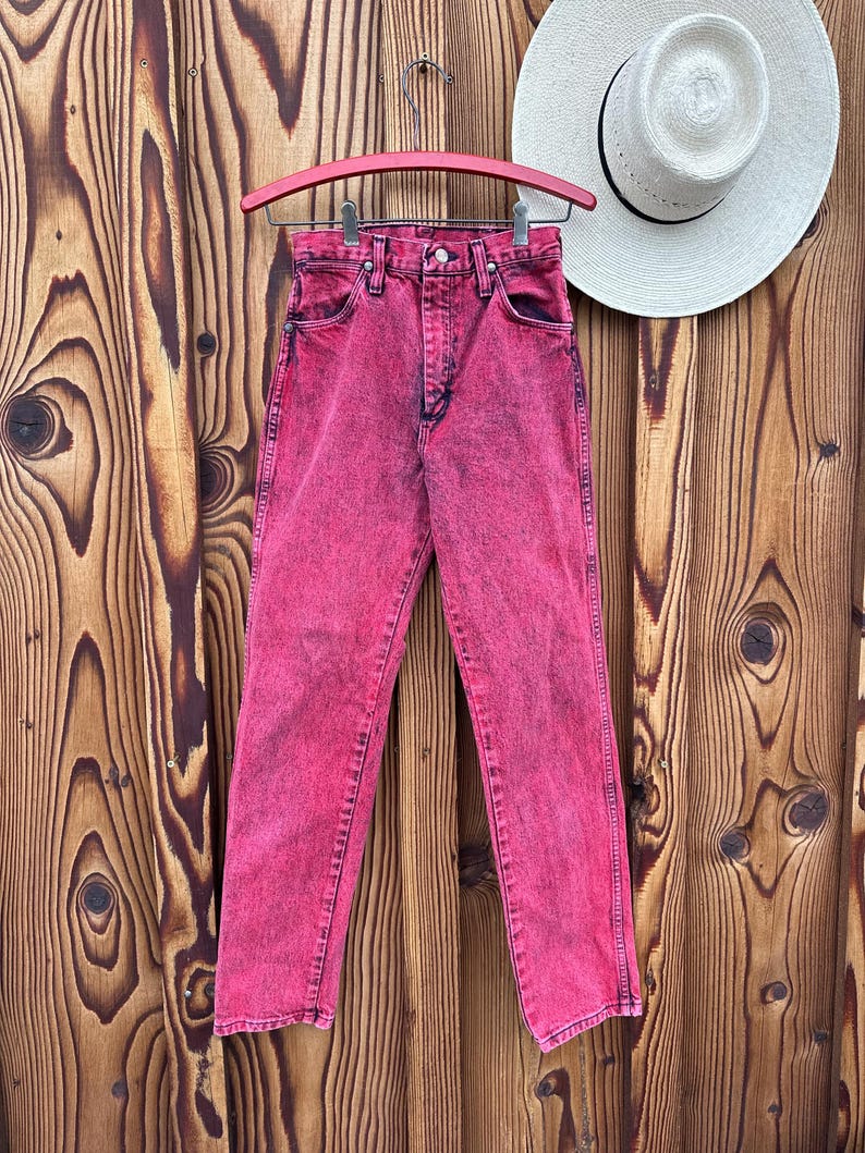 May include: A pair of acid-wash pink denim jeans hangs on a pink hanger against a wooden background. The jeans have a classic five-pocket design and a button and zipper closure. A white straw hat with a black band rests on the wooden surface.