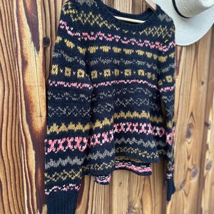 May include: A navy blue sweater with a patterned design in pink, gold, and gray. The round-neck sweater is hanging on a wooden hanger against a wooden background. A white hat is visible in the background.