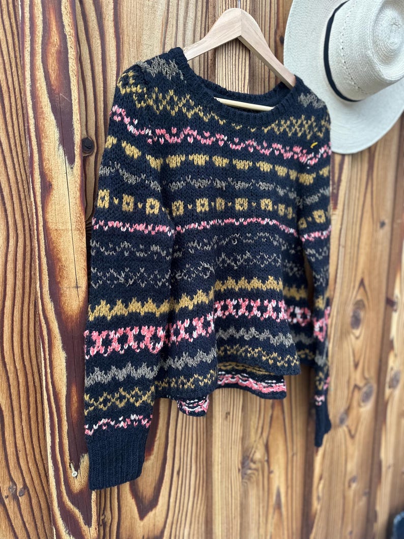 May include: A navy blue sweater with a patterned design in pink, gold, and gray. The round-neck sweater is hanging on a wooden hanger against a wooden background. A white hat is visible in the background.