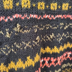 May include: Close-up of a dark navy blue knit sweater with a textured pattern. The sweater features bands of mustard yellow, coral, and tan designs. The knitwear has a cozy, handmade appearance.