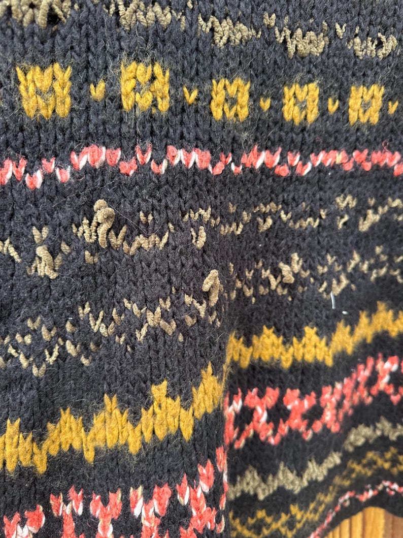 May include: Close-up of a dark navy blue knit sweater with a textured pattern. The sweater features bands of mustard yellow, coral, and tan designs. The knitwear has a cozy, handmade appearance.