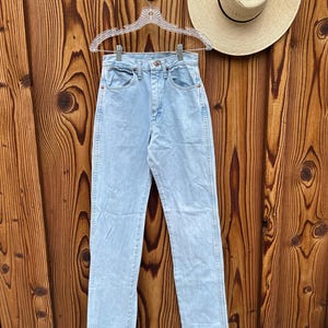 May include: Pale blue denim jeans hanging on a clear hanger against a wooden backdrop. The jeans feature a classic five-pocket design and a straight leg fit. A straw hat with a black band is also visible.