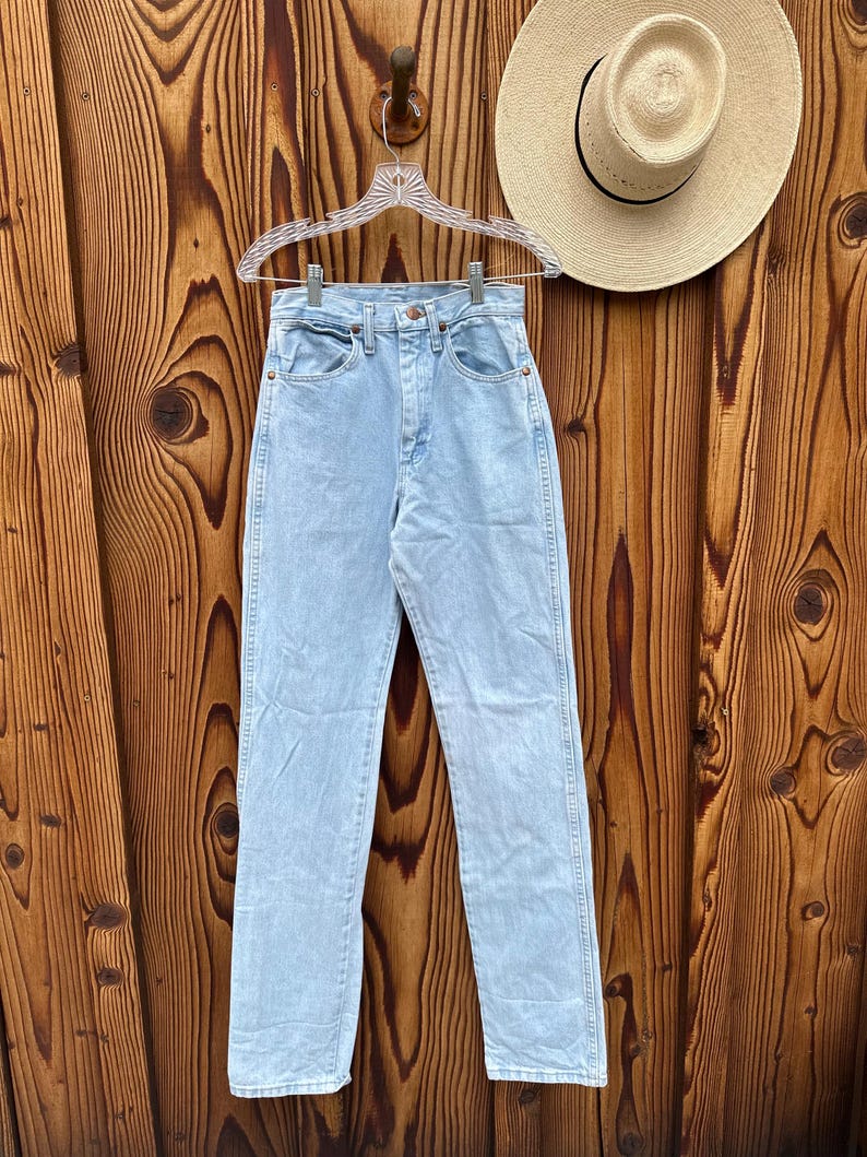 May include: Pale blue denim jeans hanging on a clear hanger against a wooden backdrop. The jeans feature a classic five-pocket design and a straight leg fit. A straw hat with a black band is also visible.