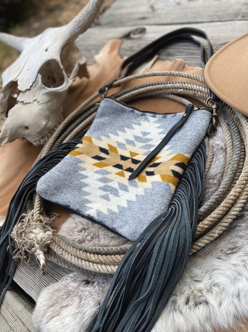 Pode incluir: Uma bolsa de ombro cinza e amarela com padr&atilde;o, com franjas pretas e uma al&ccedil;a de couro preta. A bolsa est&aacute; sobre uma superf&iacute;cie de madeira marrom com uma corda e um tapete de pele.