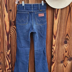 Puede incluir: Un par de pantalones vaqueros de mezclilla azul oscuro con una abertura de pierna ancha. Los pantalones tienen un estilo cl&aacute;sico de cinco bolsillos y un parche de cuero en la parte posterior con el logotipo de Wrangler.
