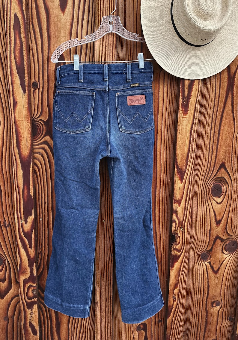 Puede incluir: Un par de pantalones vaqueros de mezclilla azul oscuro con una abertura de pierna ancha. Los pantalones tienen un estilo cl&aacute;sico de cinco bolsillos y un parche de cuero en la parte posterior con el logotipo de Wrangler.