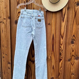 May include: Pale blue denim jeans hanging on a clear hanger. The jeans feature a classic five-pocket design with the Wrangler logo on the back pockets. A straw hat with a black band is also visible. The background is a wooden wall.