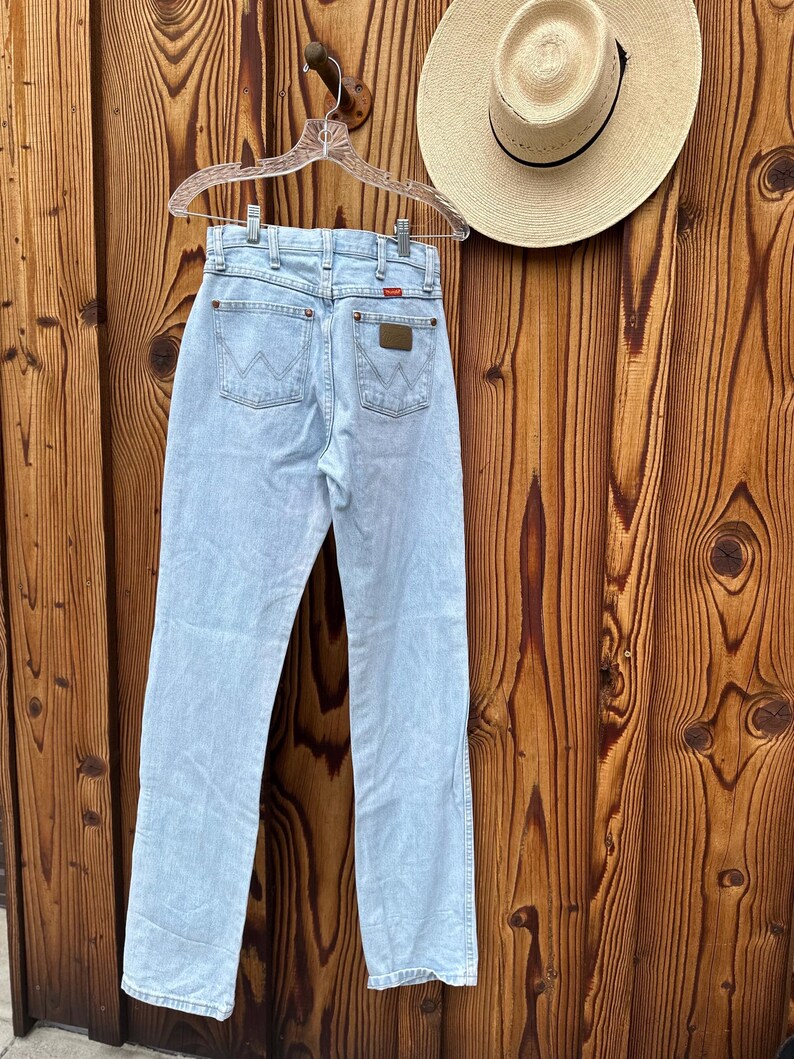 May include: Pale blue denim jeans hanging on a clear hanger. The jeans feature a classic five-pocket design with the Wrangler logo on the back pockets. A straw hat with a black band is also visible. The background is a wooden wall.
