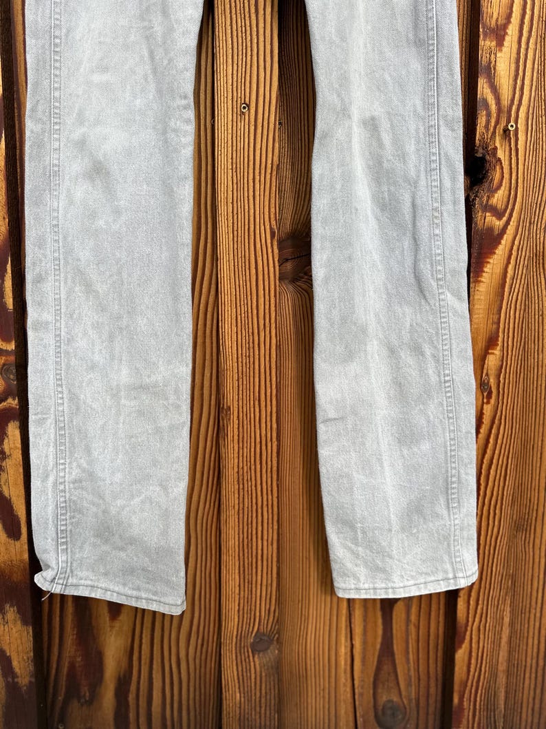 Pu&ograve; includere: Jeans in denim grigio chiaro con taglio dritto. I jeans hanno un classico design a cinque tasche e una chiusura con bottone e cerniera. Il tessuto sembra essere prelavato, conferendogli un aspetto morbido. I jeans sono esposti su uno sfondo di legno.