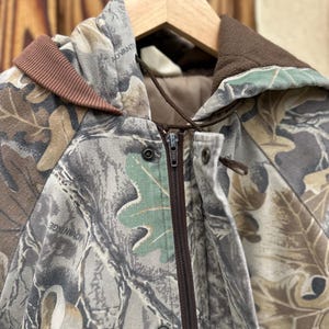 May include: A camouflage jacket with a brown zipper and a brown collar. The jacket features a leaf pattern in shades of green, brown, and gray. The word "ADVANTAGE" is visible on the jacket.