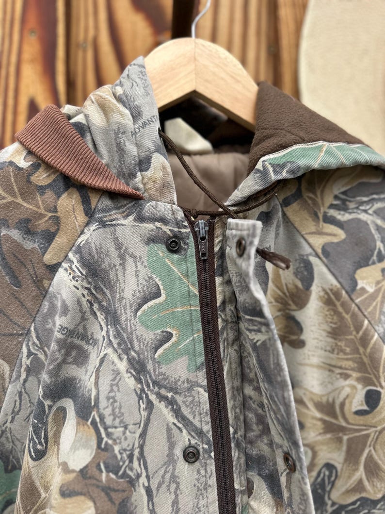 May include: A camouflage jacket with a brown zipper and a brown collar. The jacket features a leaf pattern in shades of green, brown, and gray. The word "ADVANTAGE" is visible on the jacket.