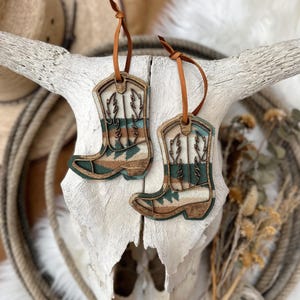 Puede incluir: Dos adornos de madera con forma de bota de vaquero con correas de cuero marrón. Las botas presentan un diseño detallado con colores turquesa, marrón y crema. Cuelgan de un cráneo blanco, con una cuerda y plantas secas en el fondo.