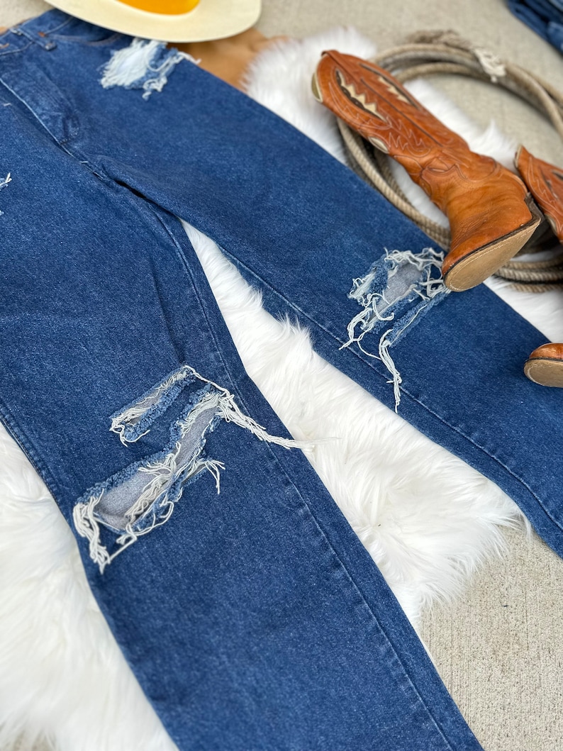 K&ouml;nnte beinhalten: Eine blaue Jeanshose mit mehreren ausgefransten L&ouml;chern und ausgefransten Kanten. Die Jeans liegt auf einer wei&szlig;en, flauschigen Oberfl&auml;che.