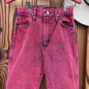 May include: A pair of acid-wash pink denim jeans hanging on a red hanger. The jeans have a classic five-pocket design with a button and zipper closure. The denim has a faded, distressed look.